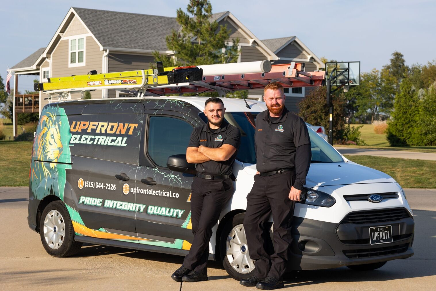Two electricians in uniform stand next to a branded Upfront Electrical van with ladders on top, parked in a residential neighborhood driveway.