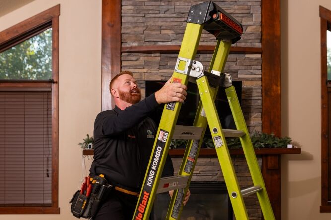 A man in work attire sets up a green ladder inside a living room, preparing to work near a fireplace and stone wall.