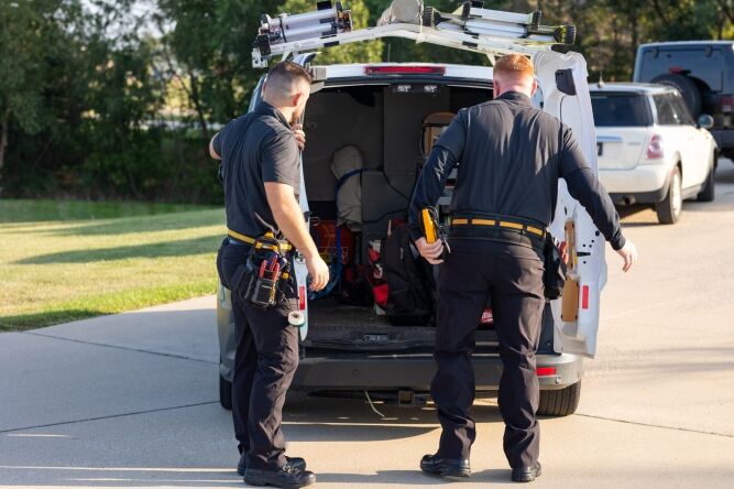 Two workers in uniforms unload tools and equipment from the back of a white utility van parked on a paved driveway.