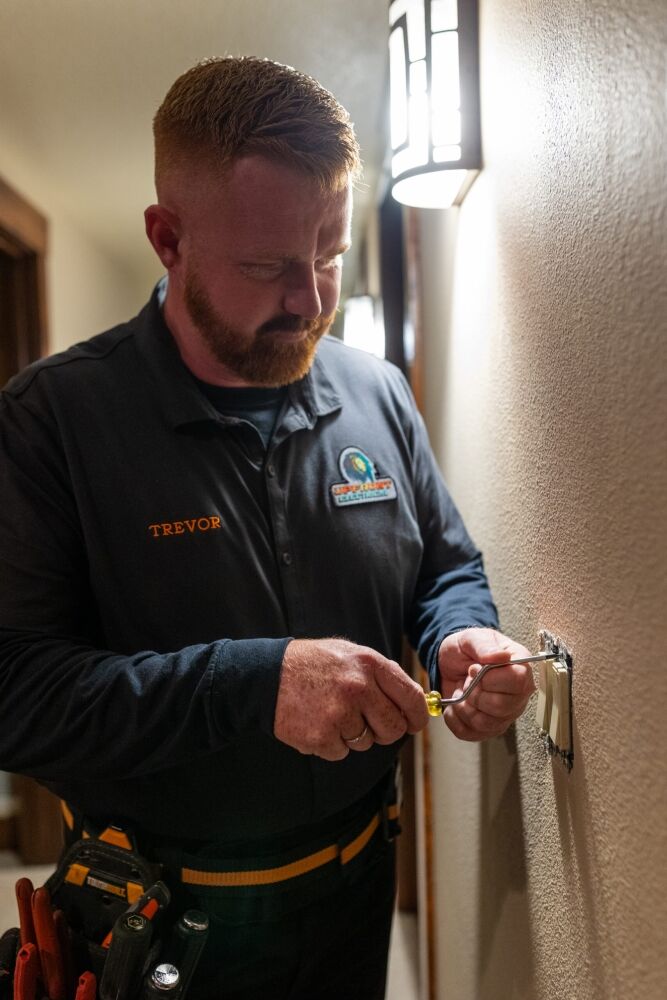 A man in a work uniform uses a screwdriver to repair or install a light switch on a wall in a well-lit hallway.