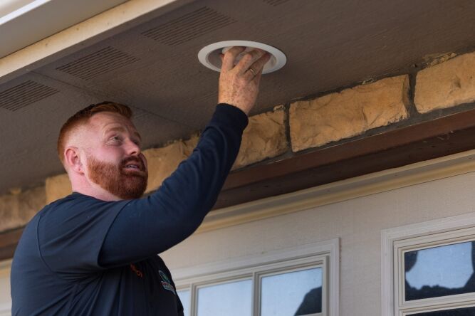 A man installs or inspects a round vent or light fixture on the ceiling of a porch near a front door.