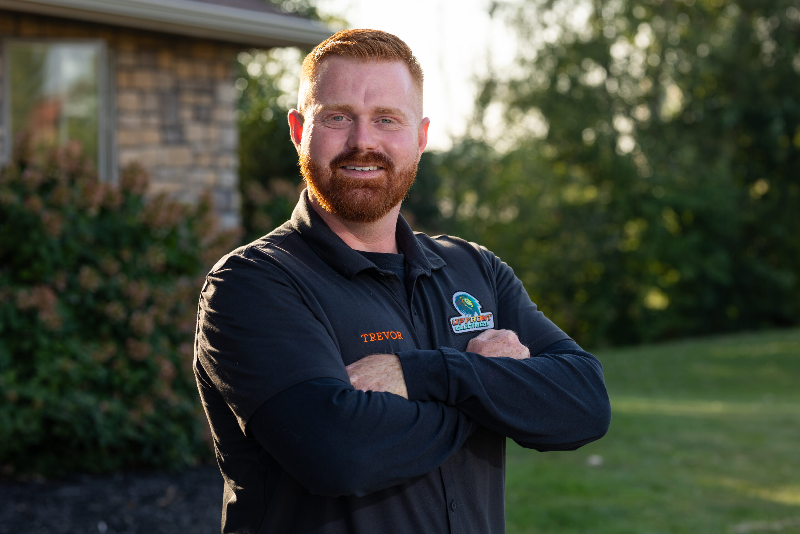 A man with red hair and beard stands outdoors with arms crossed, wearing a black collared shirt embroidered with TREVOR and a colorful logo.