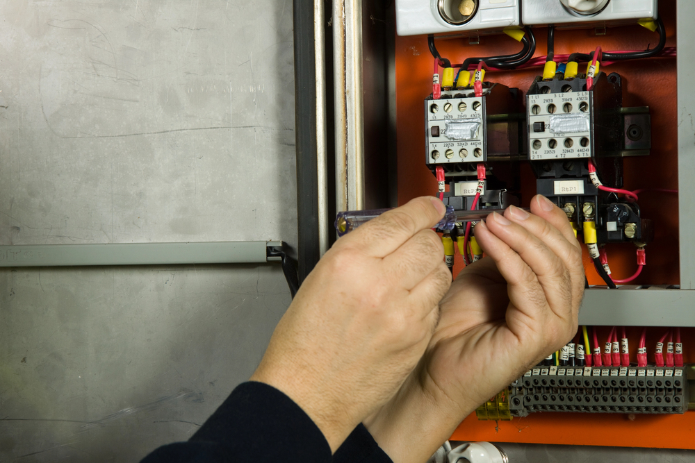 Hands using a screwdriver to adjust wiring inside an open electrical control panel, demonstrating the importance of electrical safety inspections and code corrections among various visible wires and components.