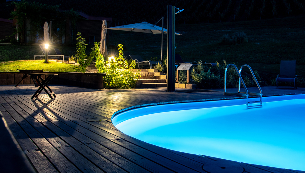 A swimming pool glows with blue lights at night, surrounded by wooden decking, steps, and outdoor furniture in a landscaped garden—perfect for relaxing after professional pools spas and hot tub wiring.