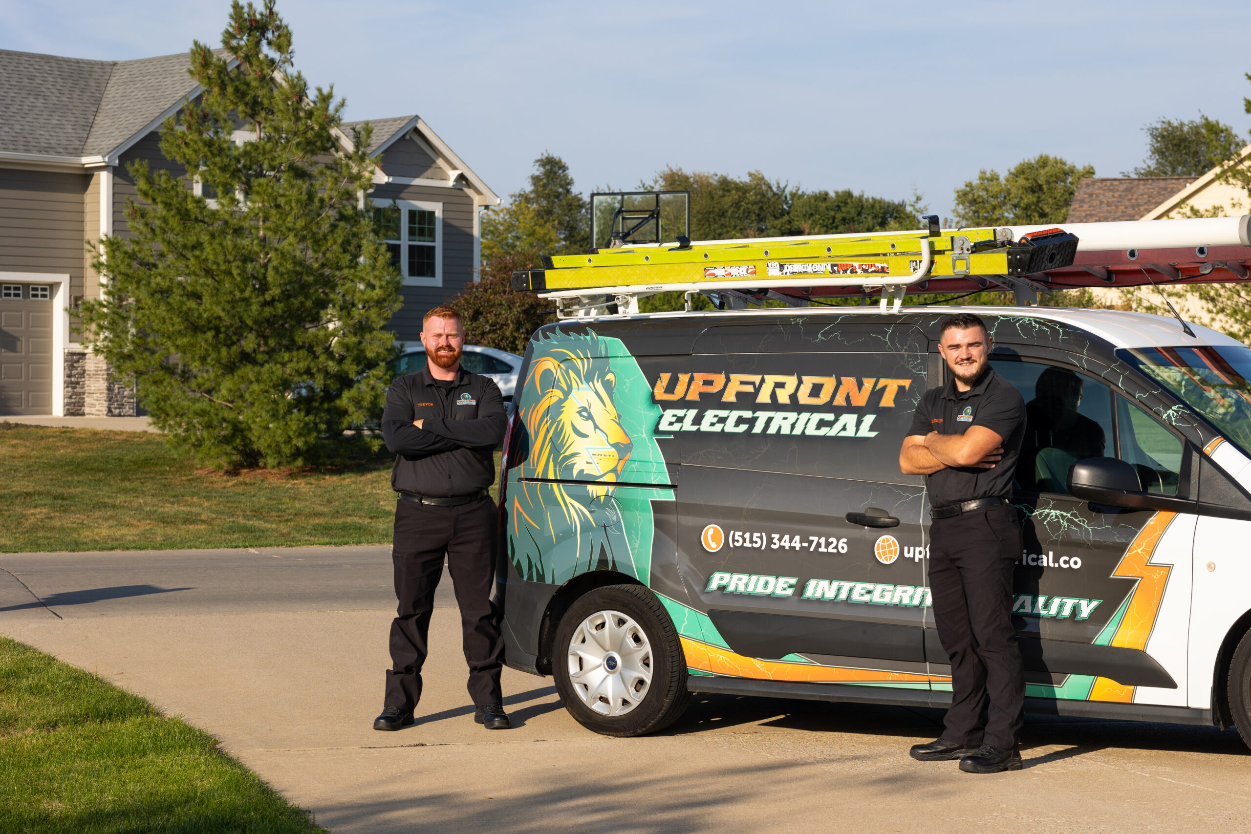 Two electricians in black uniforms stand with arms crossed next to a branded Upfront Electrical van parked on a residential street.