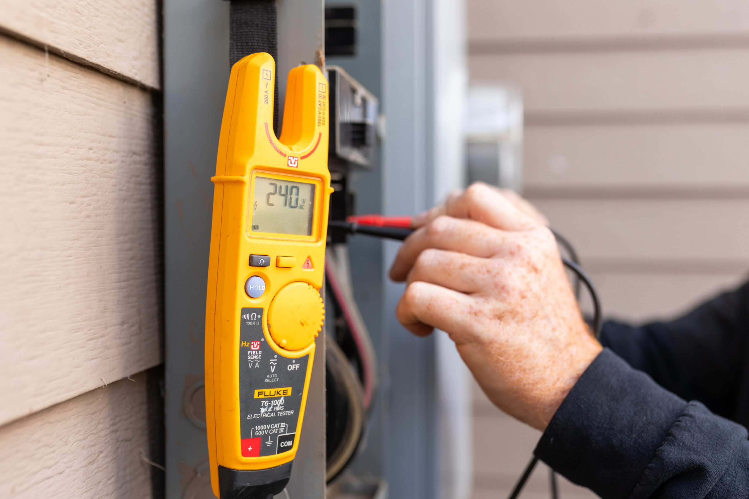 A person uses a digital clamp meter to measure voltage in an electrical panel; the display shows a reading of 240.7 volts.