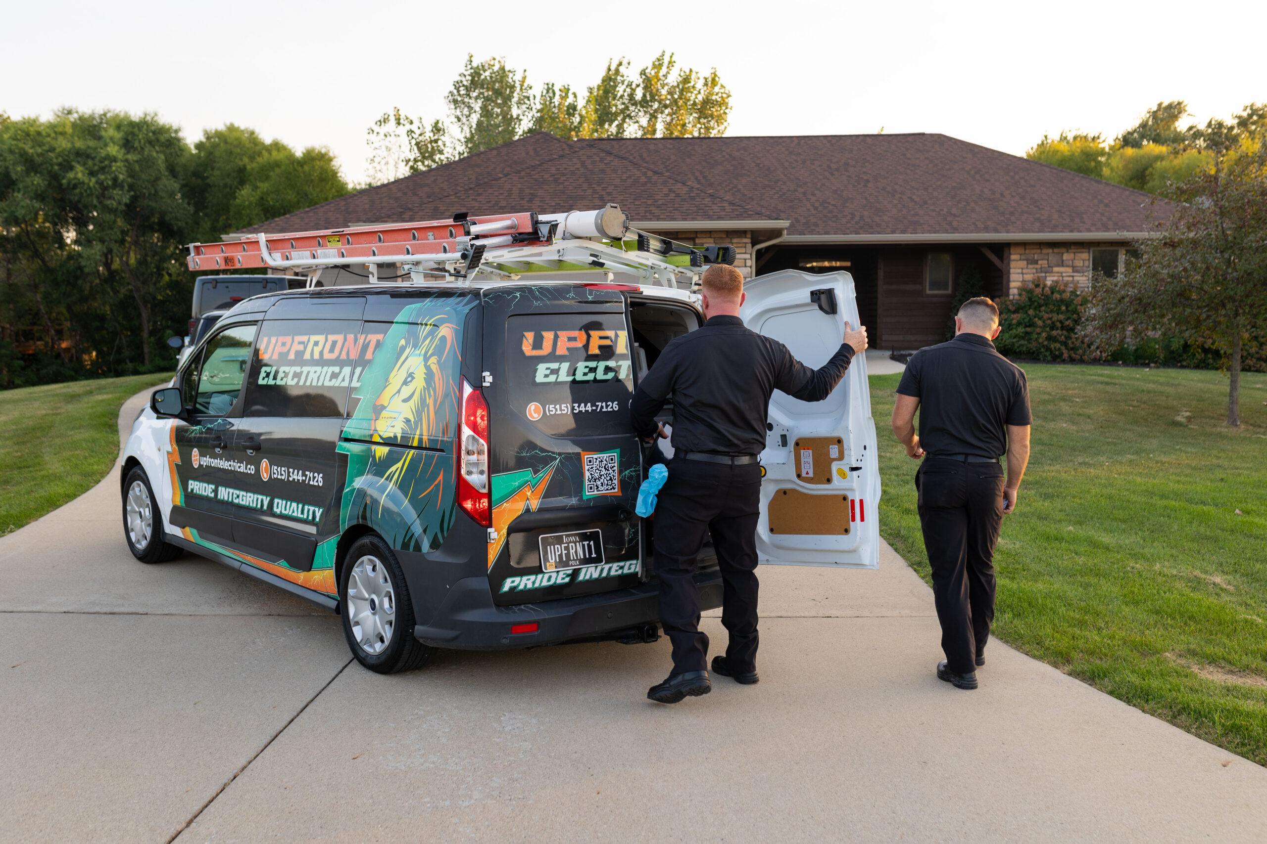 Two men in black uniforms stand next to an Upfront Electrical van parked in a driveway in front of a house, with one man opening the van's rear door.