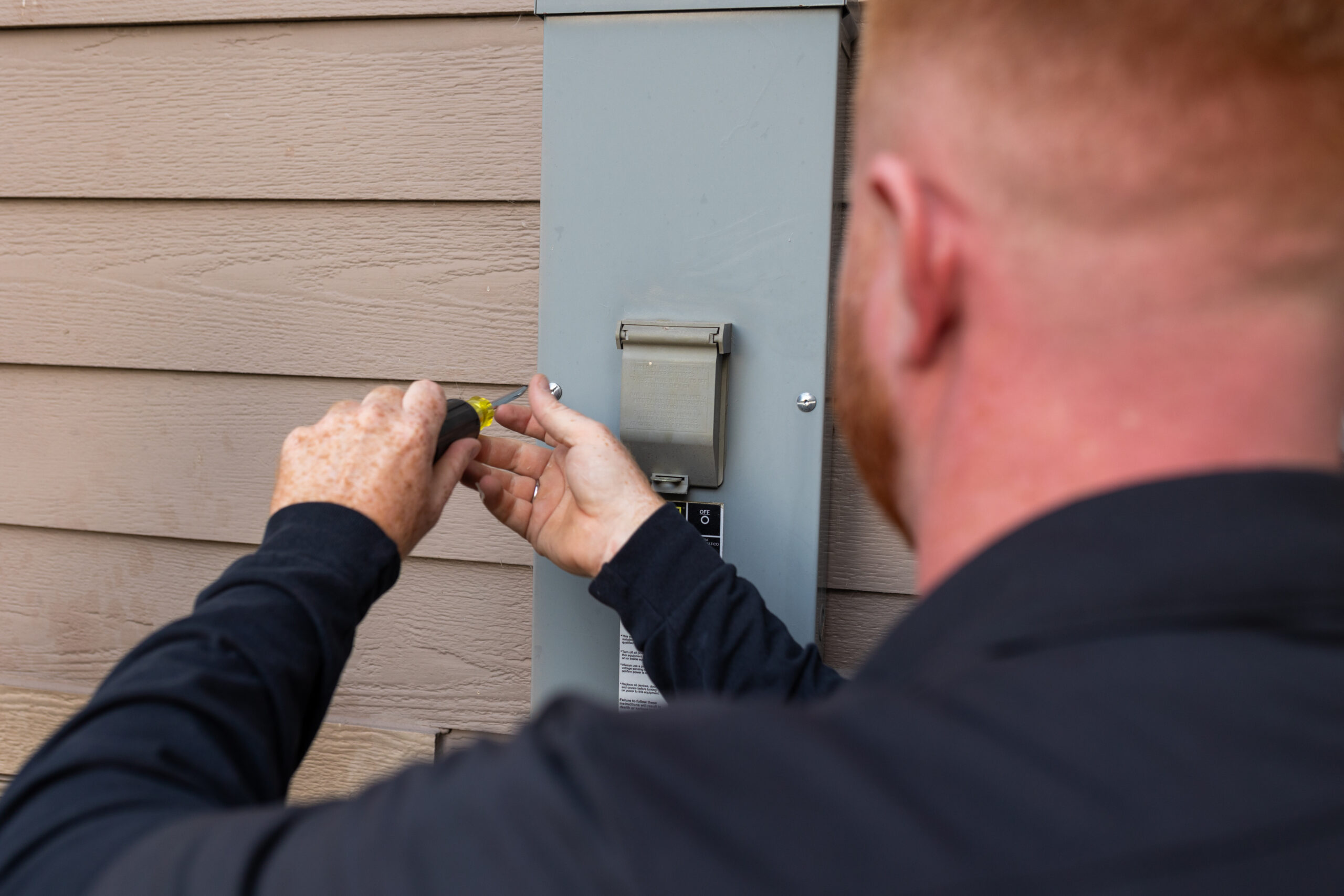 Person using a screwdriver to work on an outdoor electrical panel mounted on the side of a house.